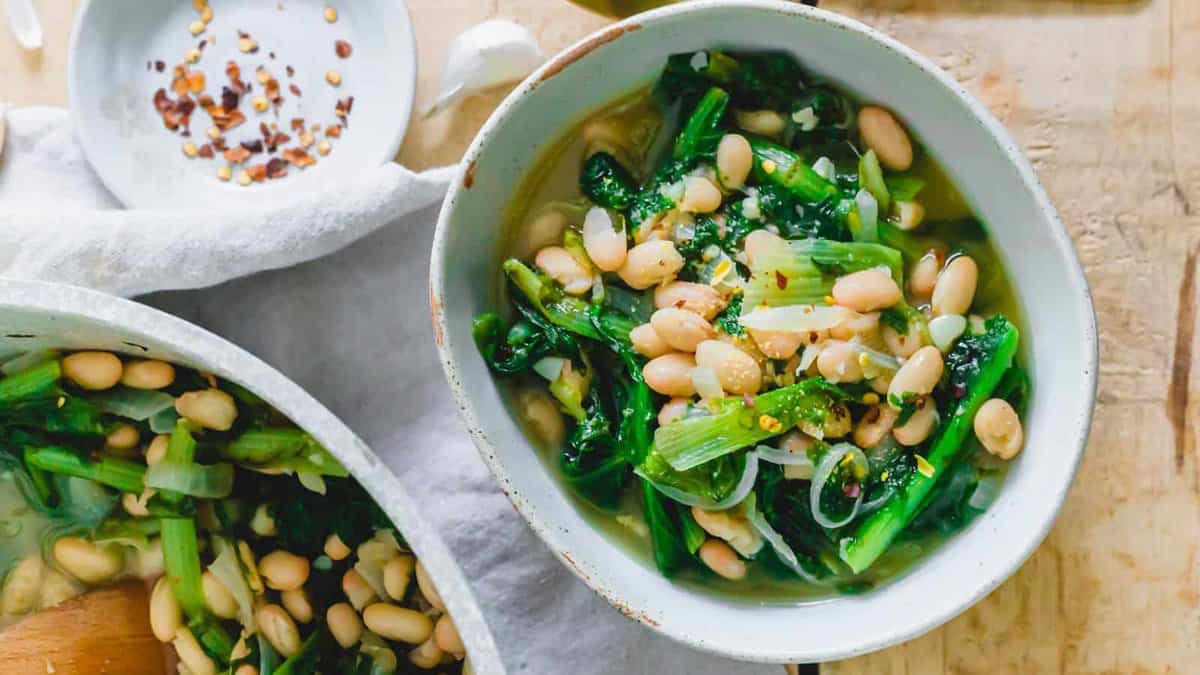 A bowl of white bean and leafy green vegetable soup garnished with red pepper flakes, with a dish of red pepper flakes and another pot of soup nearby.