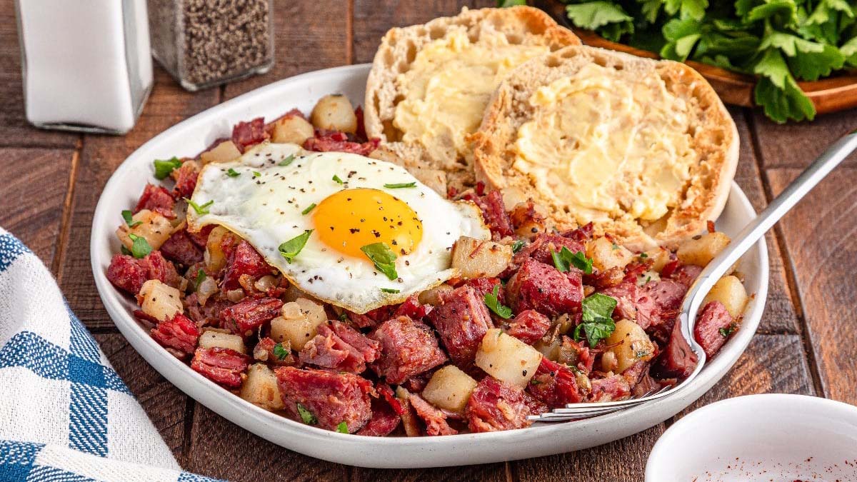 A plate of corned beef hash with diced potatoes, topped with a sunny-side-up egg, served with a buttered English muffin on the side.