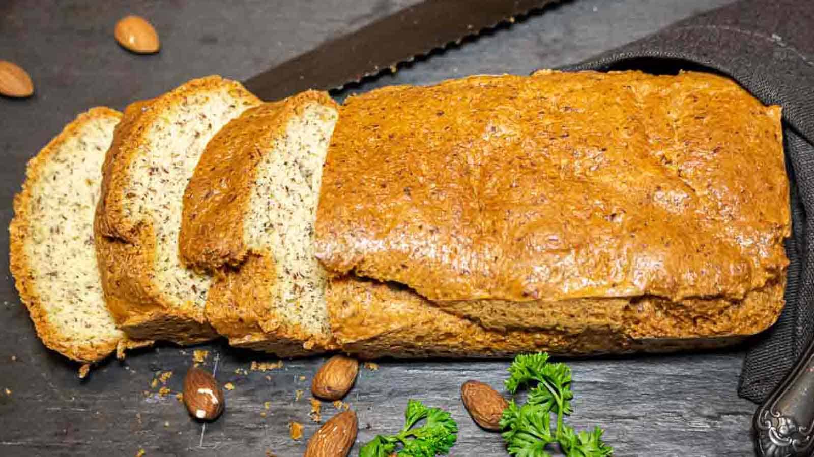 A partially sliced loaf of homemade bread sits on a dark surface with scattered whole almonds and sprigs of parsley nearby.