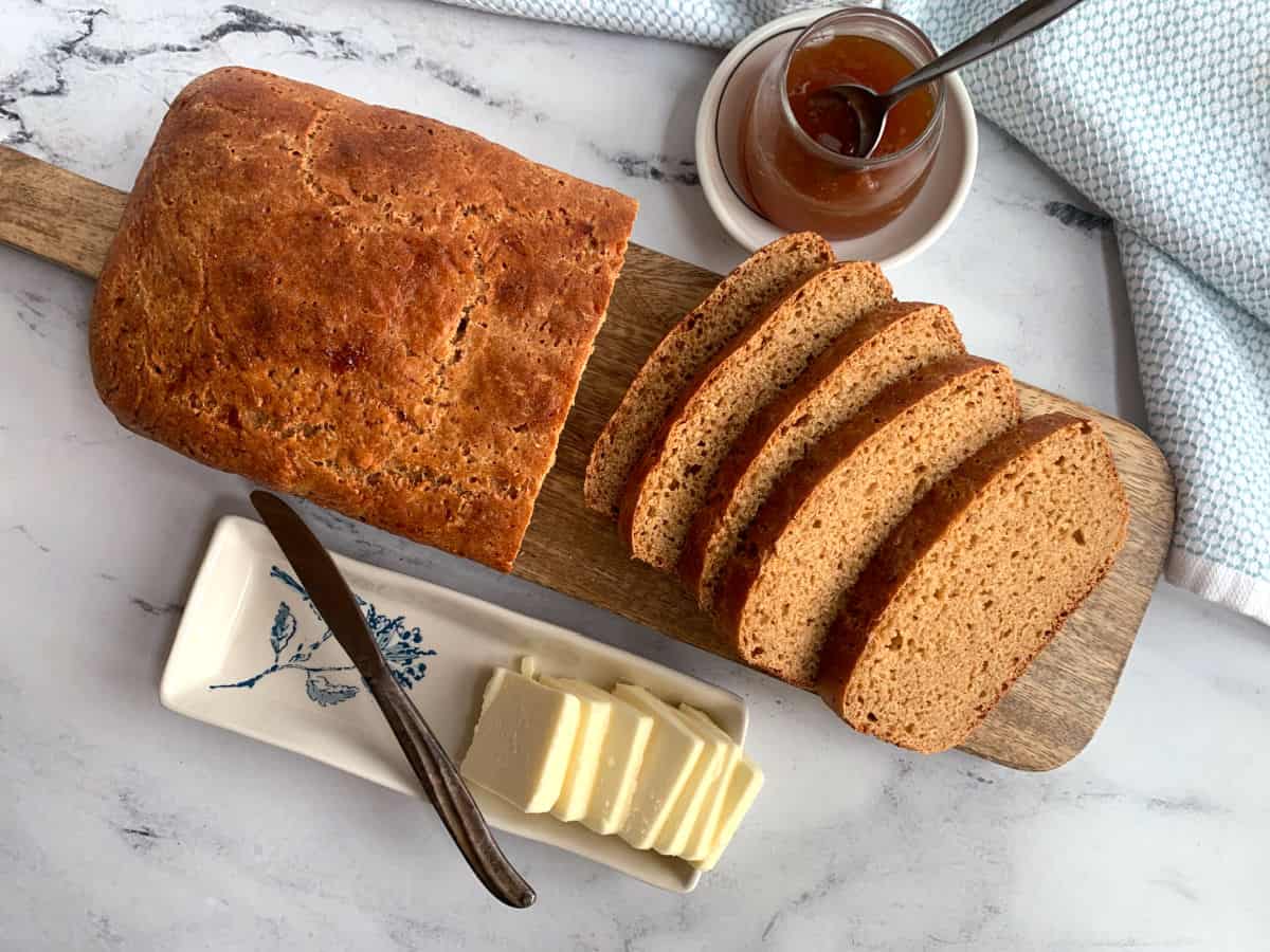 A loaf of sliced bread on a wooden board with a dish of butter, a knife, and a jar of jam on a marble surface.