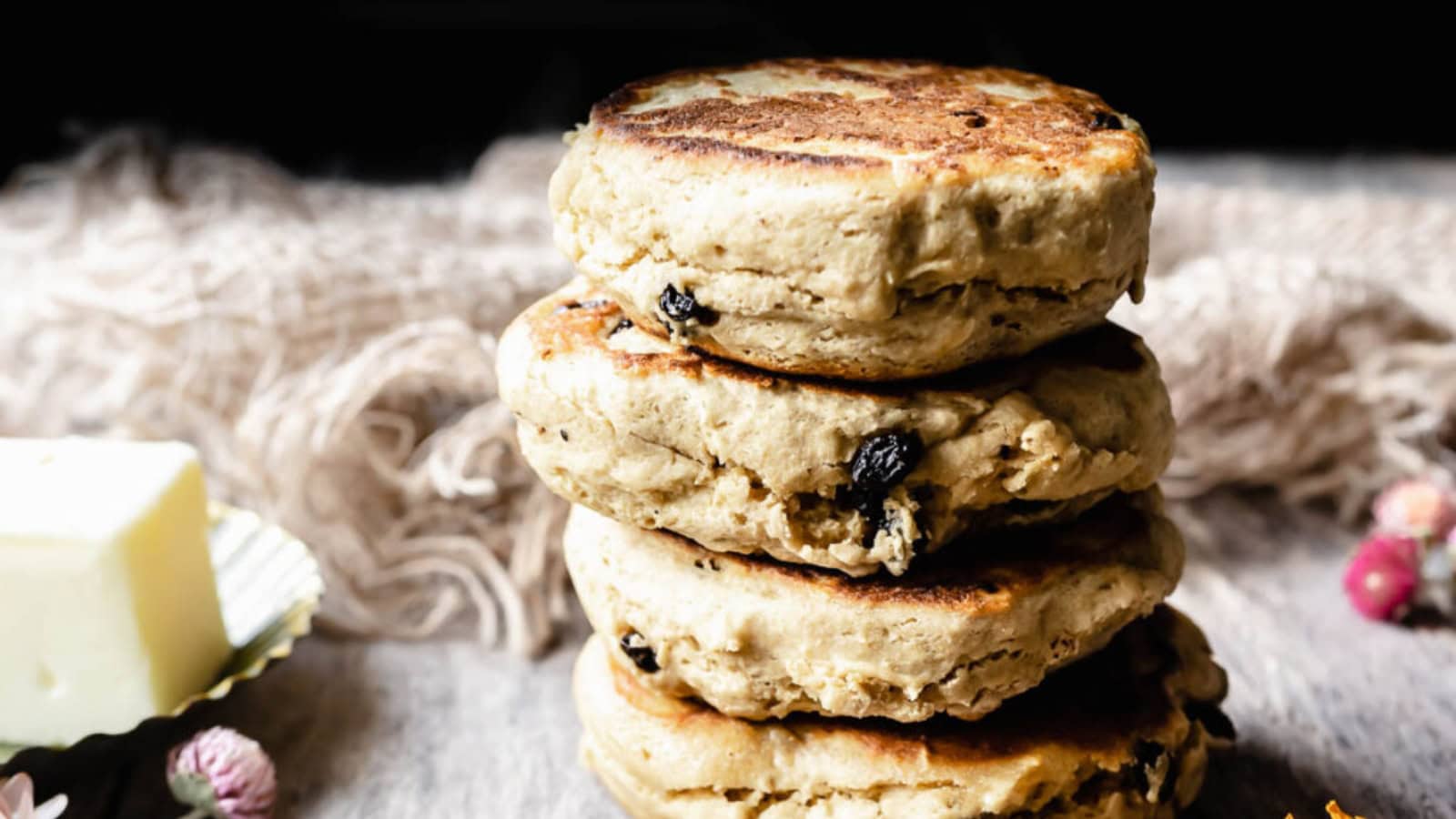 Four golden brown Welsh cakes stacked on a table, with visible raisins and a pat of butter in the background.