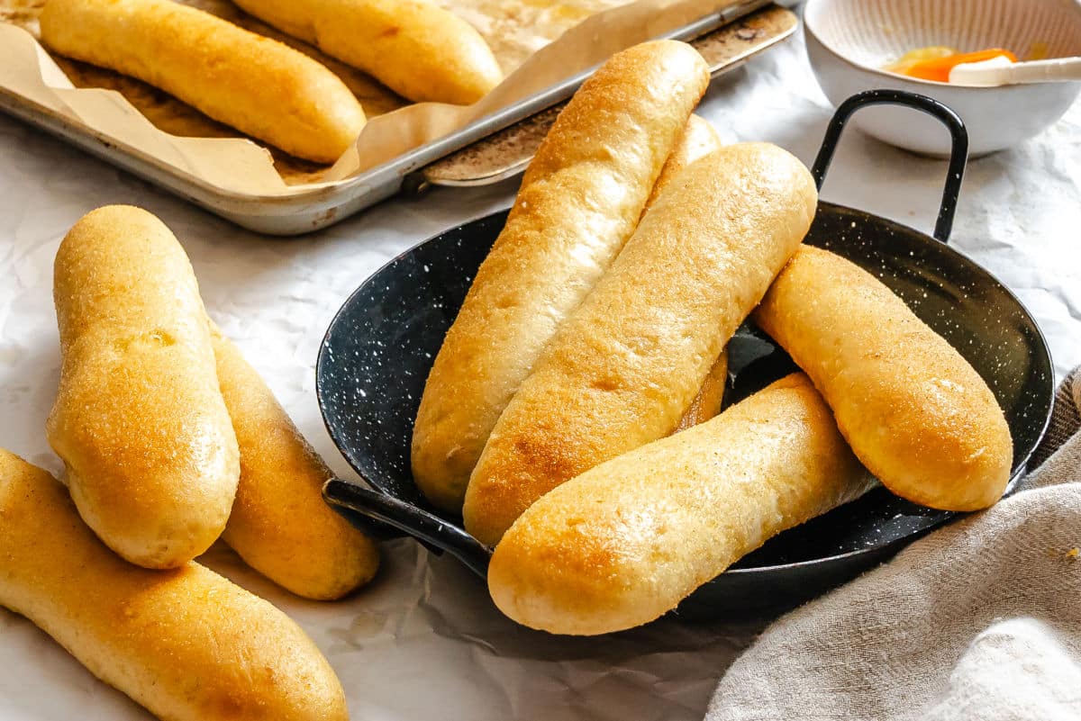 Several golden-brown breadsticks are arranged in a black pan, with more breadsticks on a baking tray and the table nearby.