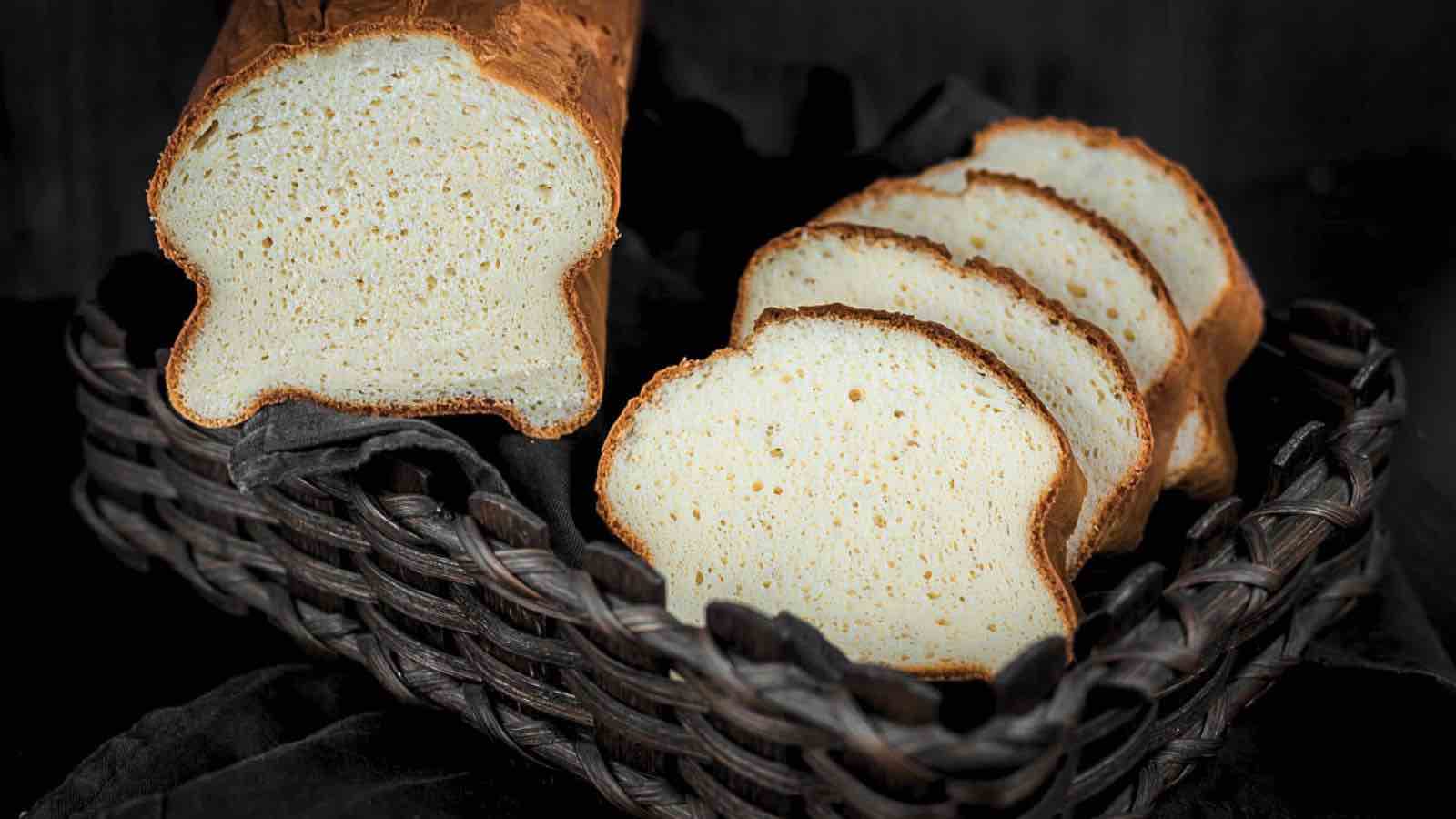 A loaf of bread with several slices cut, displayed in a woven basket on a dark cloth background.