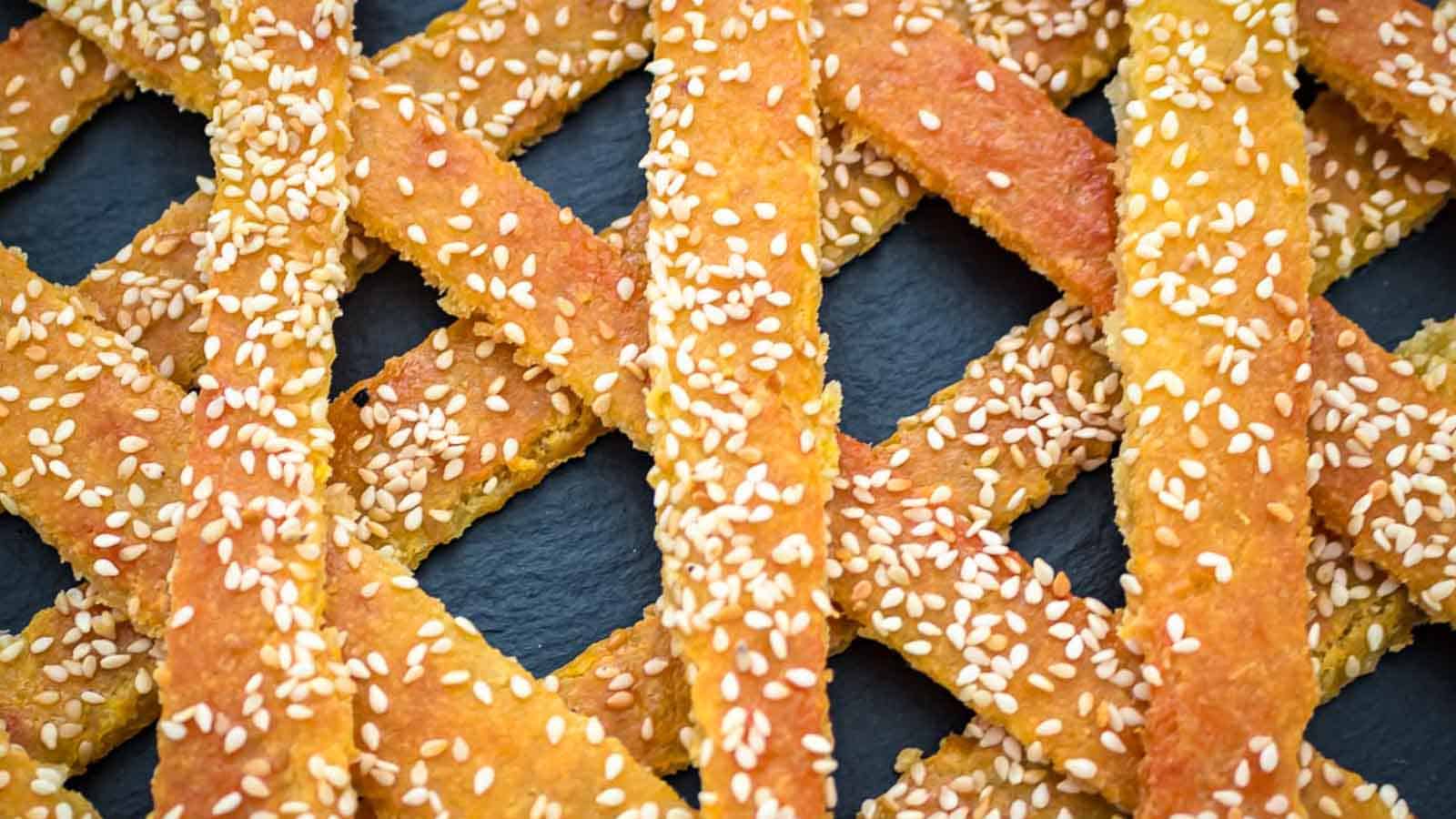 Close-up of golden, baked sesame breadsticks arranged in a crisscross pattern on a dark surface.