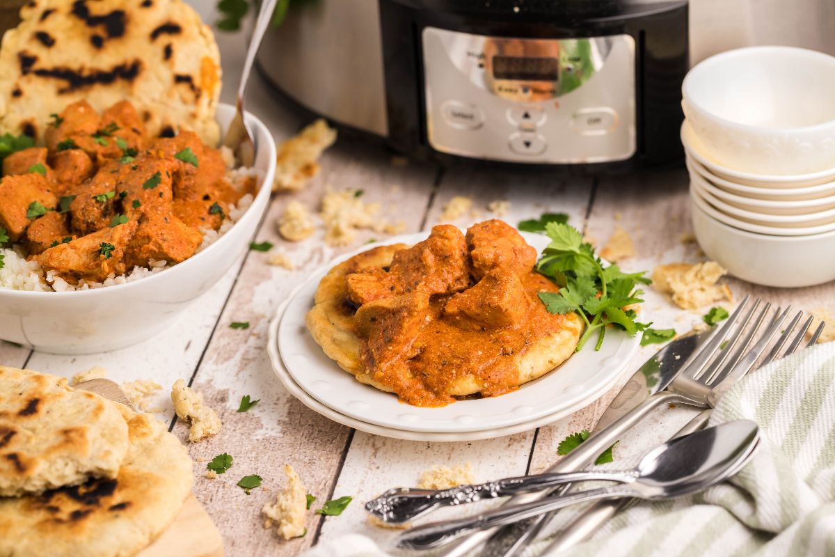 A plate of naan topped with chicken curry sits on a table near a bowl of curry over rice, utensils, stacked bowls, and a slow cooker in the background.