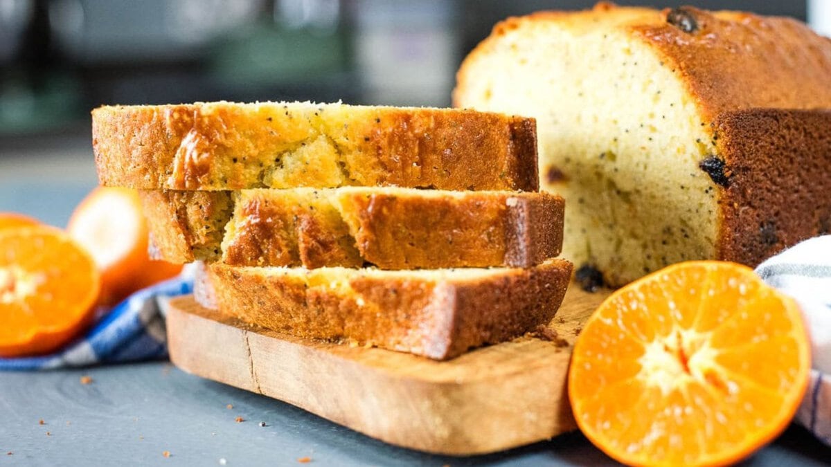 Three slices of orange loaf cake on a wooden board, with a halved mandarin orange and a whole loaf in the background.