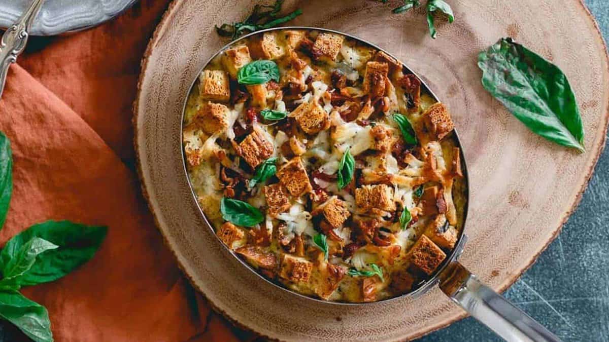 A pan of savory bread pudding topped with toasted croutons and fresh basil is placed on a wooden tray, with basil leaves scattered around.