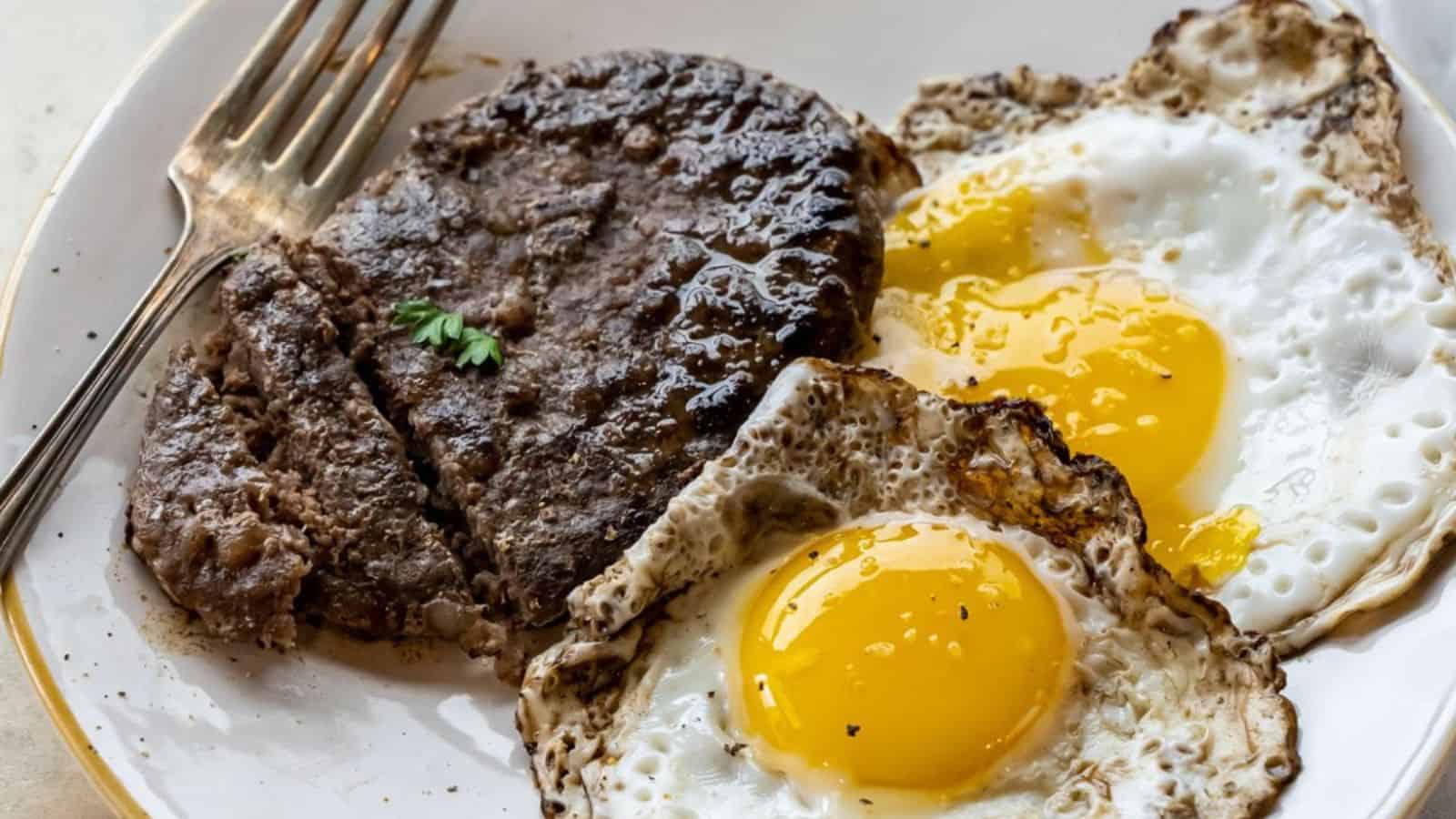 A plate with two sunny-side-up eggs and a cooked hamburger patty, with a fork resting on the edge.