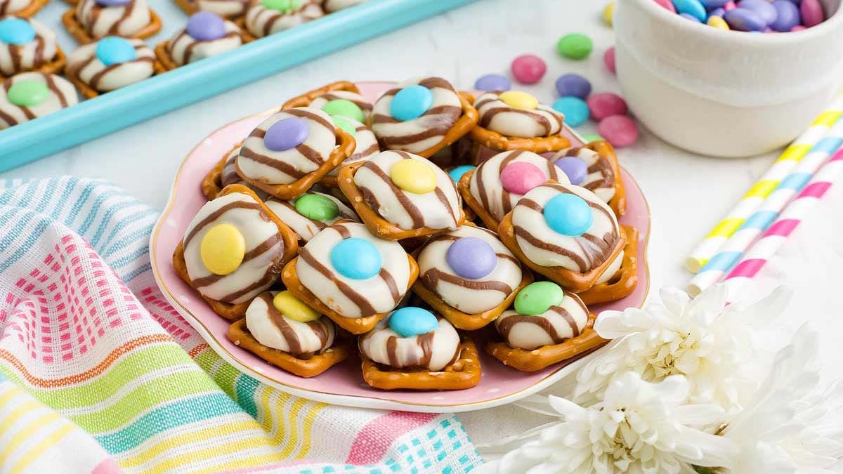 A plate of pretzel bites topped with striped chocolate, melted white chocolate, and colorful candy-coated chocolates sits on a table with a striped cloth and flowers.