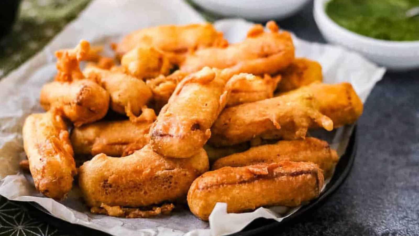 A plate of golden-brown fried banana fritters is served on parchment paper, with a bowl of green dipping sauce in the background.