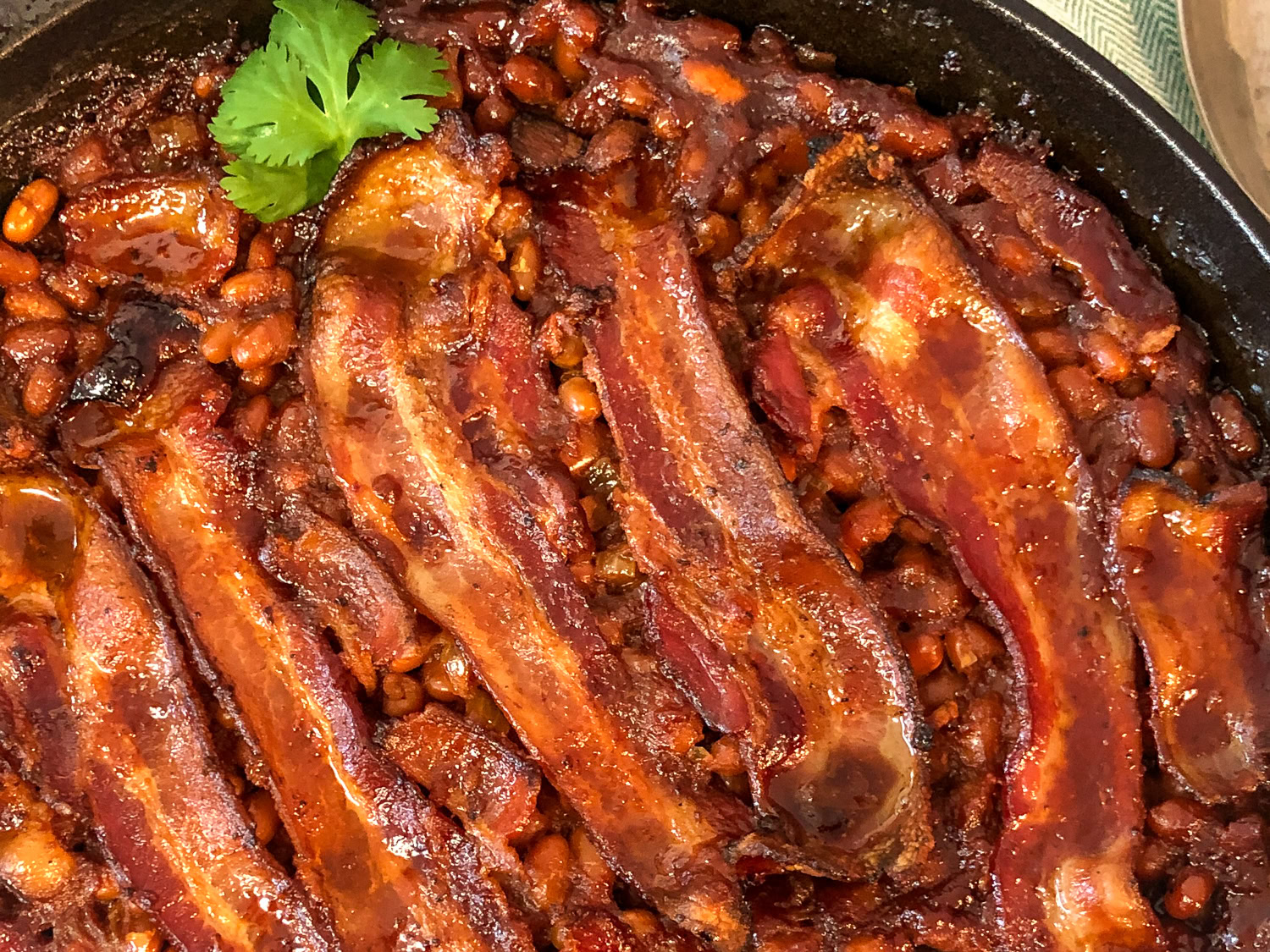 Baked beans topped with strips of cooked bacon in a skillet, garnished with a sprig of cilantro.