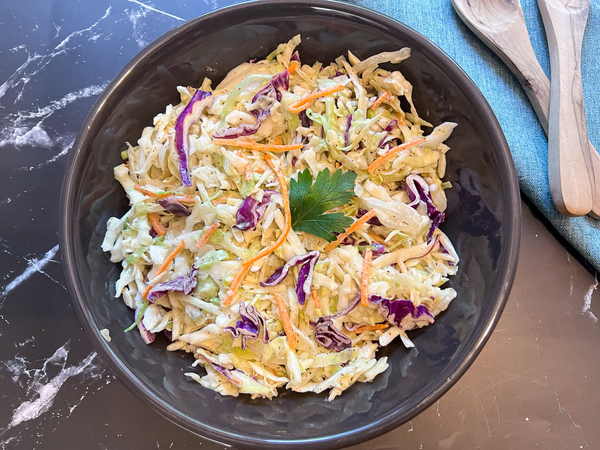 A bowl of coleslaw with shredded green and purple cabbage, carrots, and a parsley garnish, placed on a dark marble surface next to a blue napkin and wooden utensils.