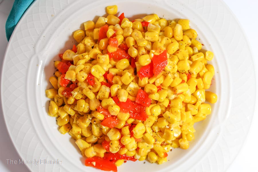 A white plate filled with cooked corn kernels and red bell pepper pieces, seasoned with black pepper.