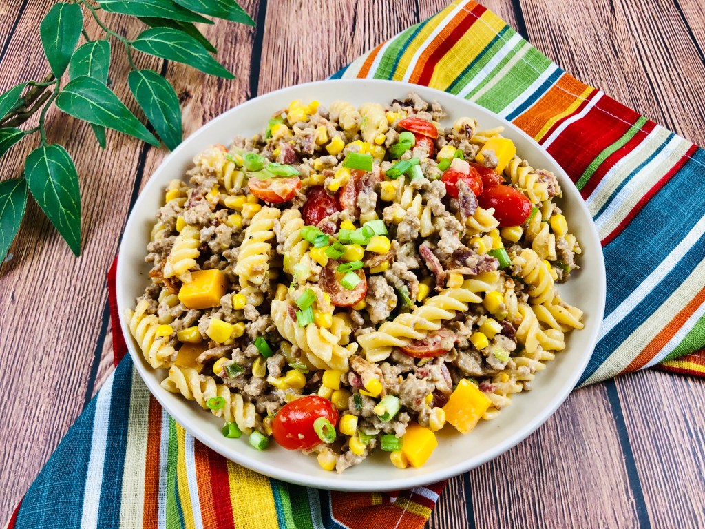 A bowl of rotini pasta salad with ground meat, corn, cherry tomatoes, diced yellow peppers, and green onions on a colorful striped cloth, set on a wooden surface.