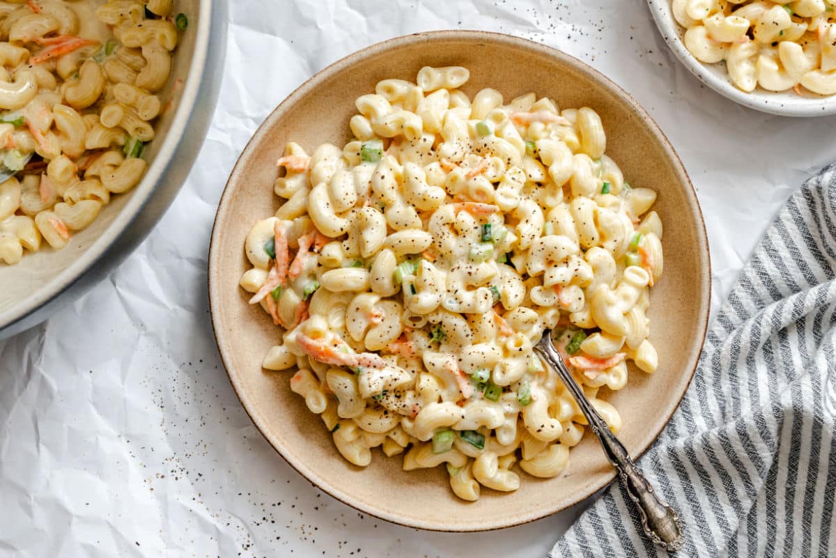 A plate of creamy macaroni salad with chopped green onions and shredded carrots, served with a fork on a beige dish.