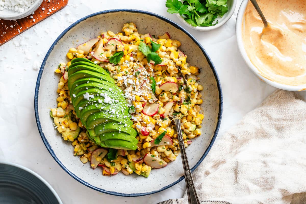 A bowl of corn salad topped with sliced avocado, radishes, crumbled cheese, and cilantro, surrounded by a small bowl of sauce, cilantro, and a napkin.