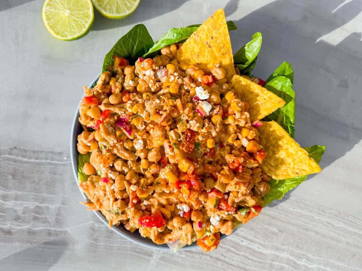 A bowl of chickpea salad with diced vegetables and herbs, garnished with three tortilla chips and green leaves, with lime slices on the side.