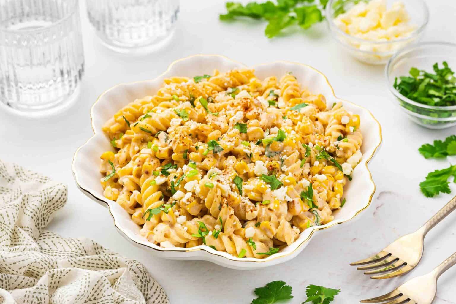A bowl of rotini pasta mixed with a creamy sauce, garnished with chopped herbs and crumbled cheese, set on a white surface with utensils and glassware nearby.
