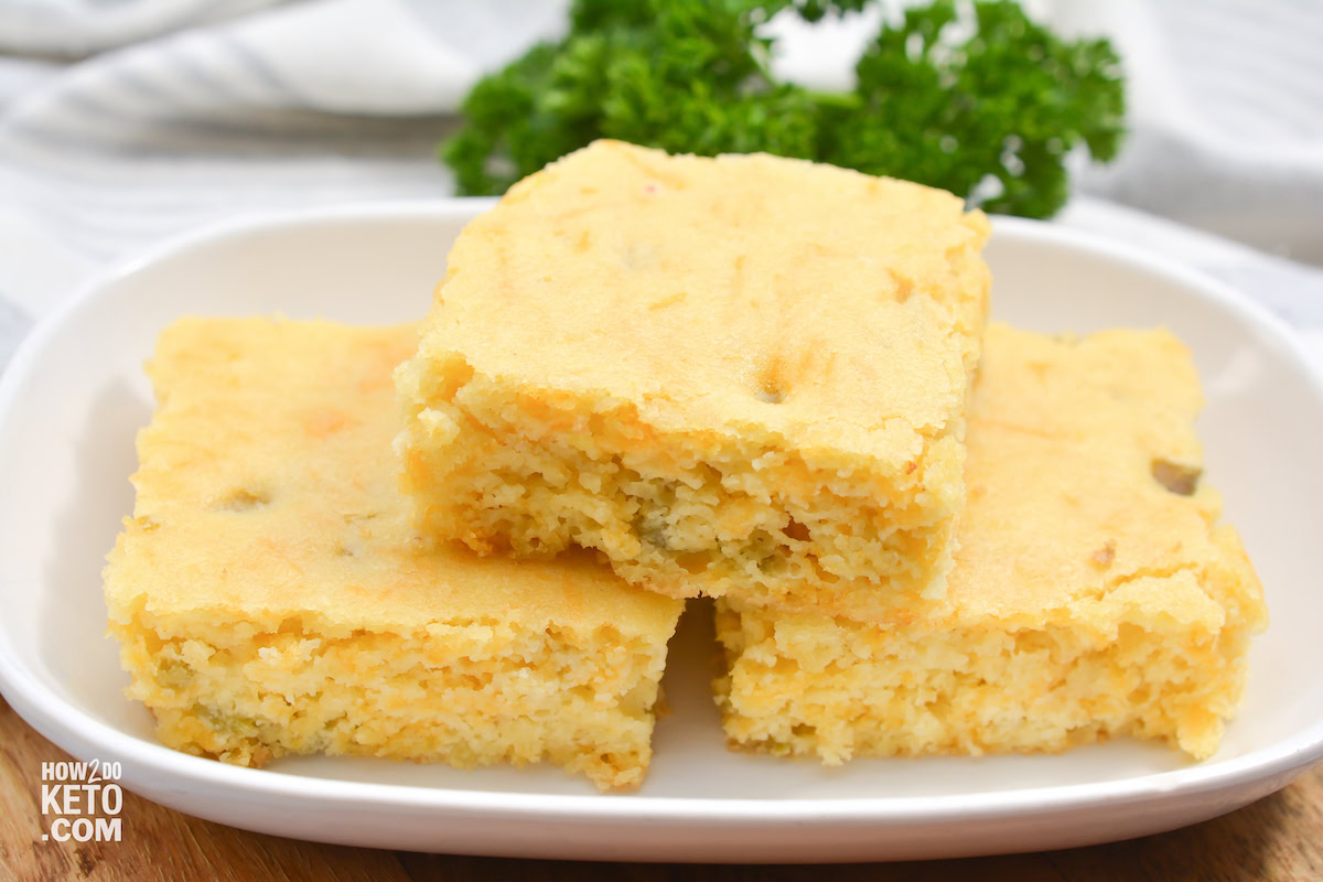 Three rectangular pieces of cornbread are stacked on a white plate, with a bunch of parsley in the background.