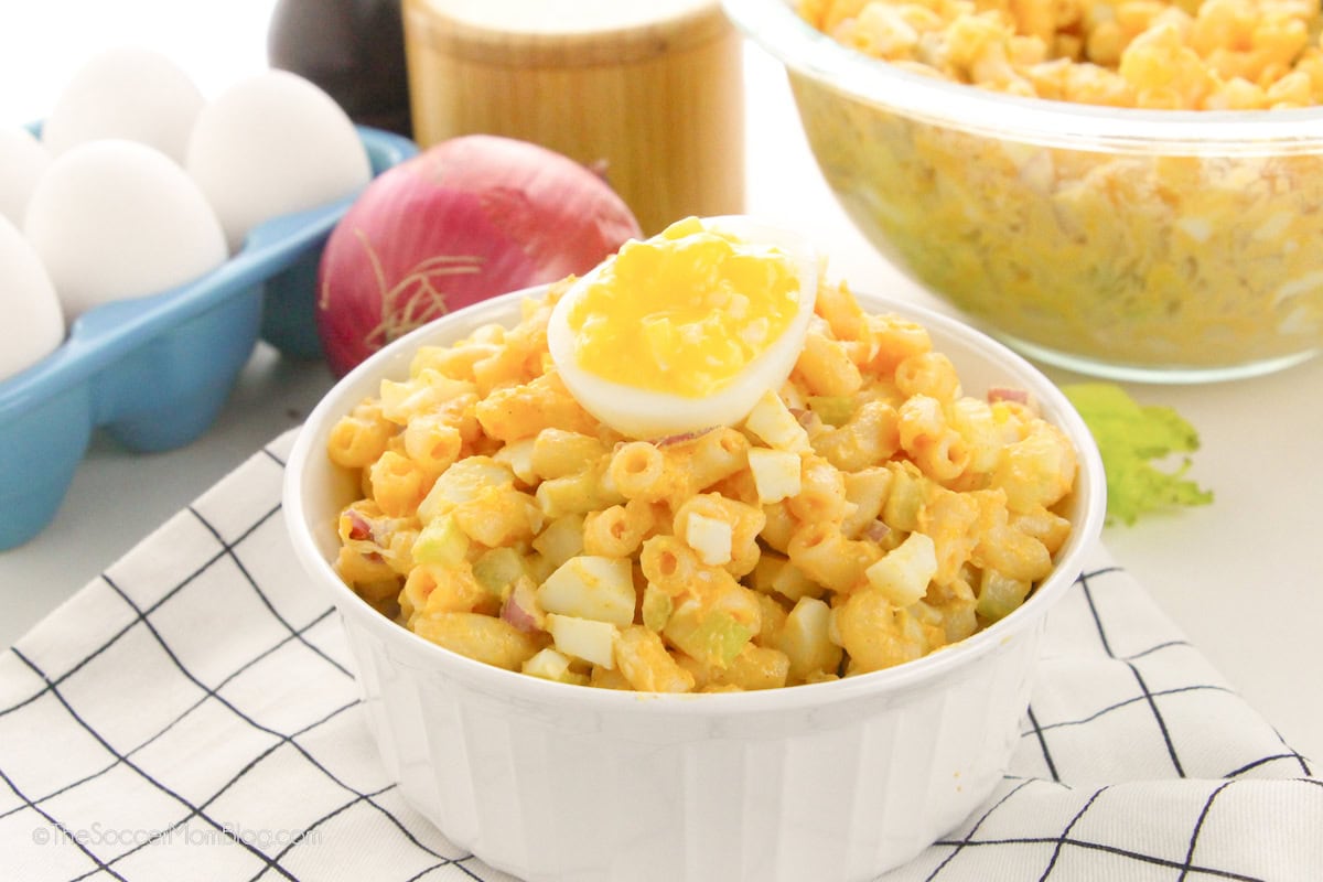 A white bowl of macaroni salad topped with a halved boiled egg, surrounded by raw eggs, a red onion, and a glass dish of macaroni salad on a checkered cloth.