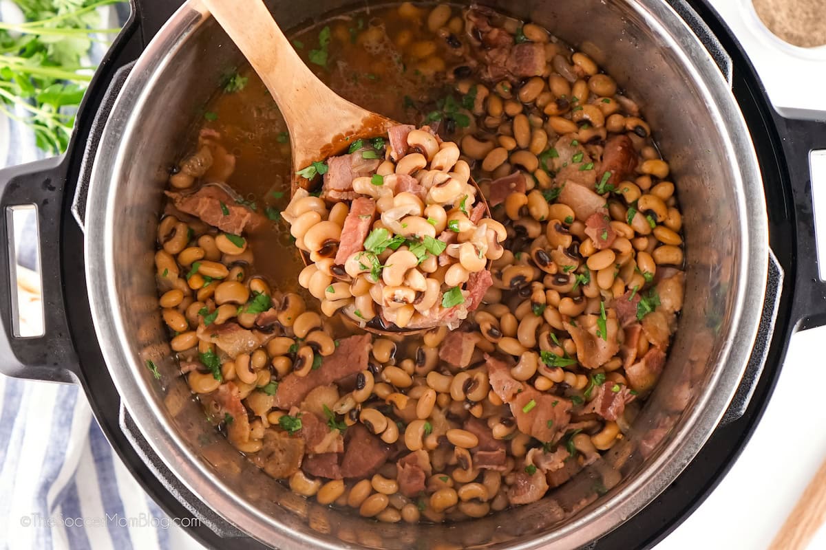 A pot of cooked black-eyed peas with pieces of ham, garnished with chopped herbs, with a wooden spoon lifting a portion from the pot.