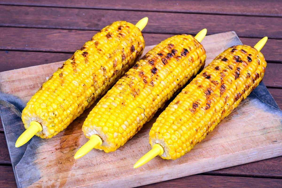 Three grilled corn on the cob with yellow corn holders placed on a wooden cutting board.
