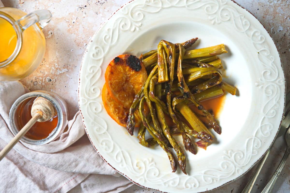 White plate with roasted asparagus and a grilled orange slice, next to a jar of honey with a dipper and a glass pitcher of orange liquid on a light surface.