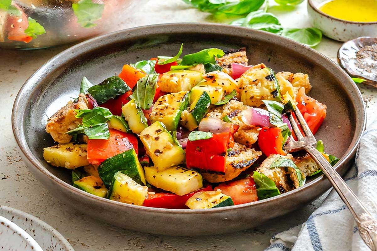 A bowl of panzanella salad with chunks of bread, zucchini, tomatoes, red onion, red bell pepper, and fresh basil leaves, served with a fork.