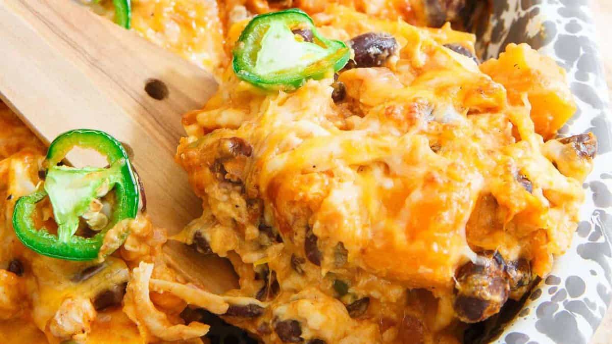 Close-up of a cheesy casserole with black beans, shredded meat, and sliced jalape&ntilde;os, being served with a wooden spatula.