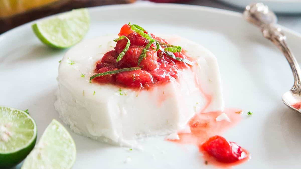 A serving of panna cotta topped with strawberry sauce and thin lime zest strips, garnished with lime wedges on a white plate.