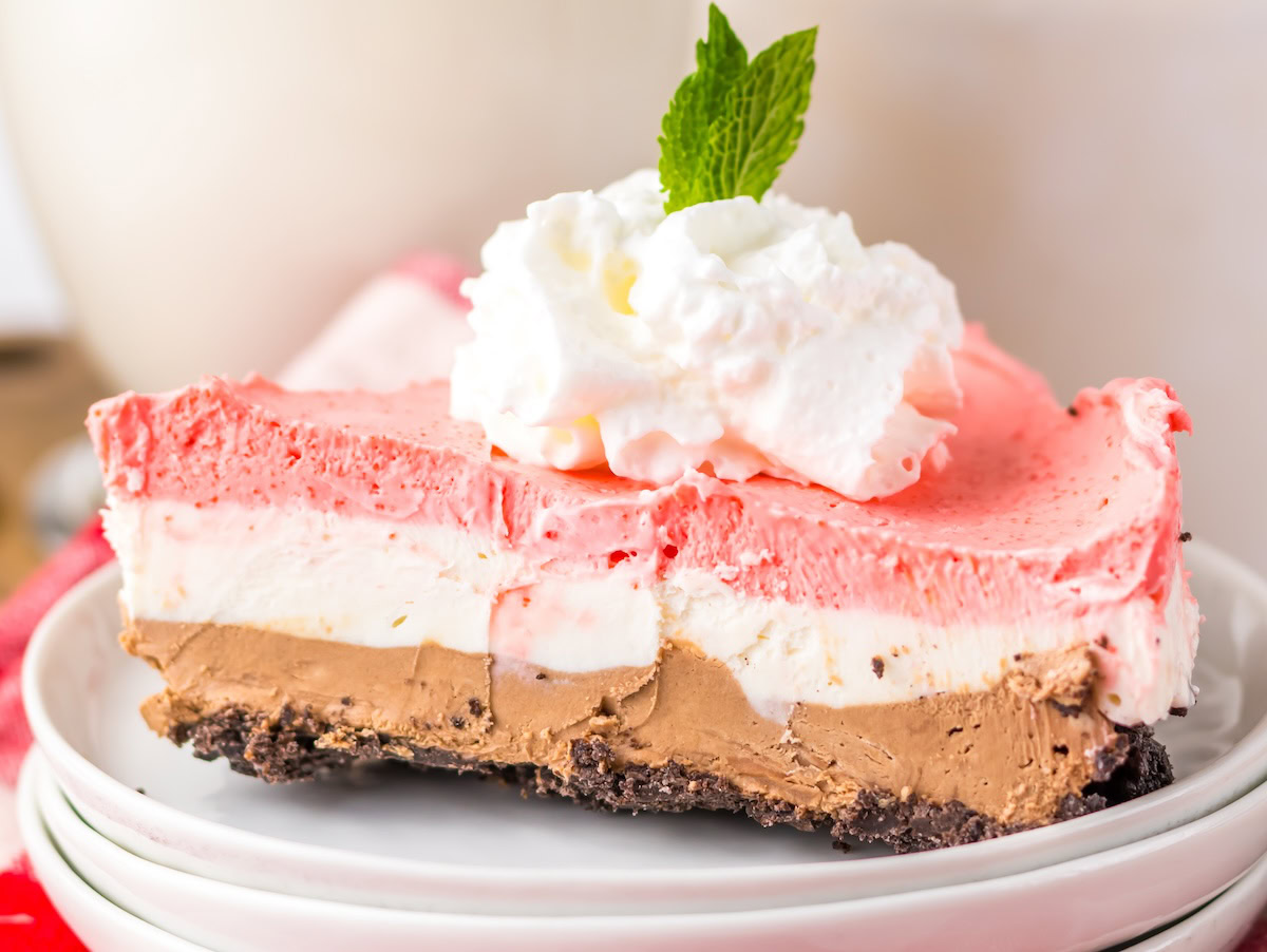 A slice of layered ice cream dessert with chocolate, vanilla, and strawberry layers on a plate, topped with whipped cream and a mint leaf.