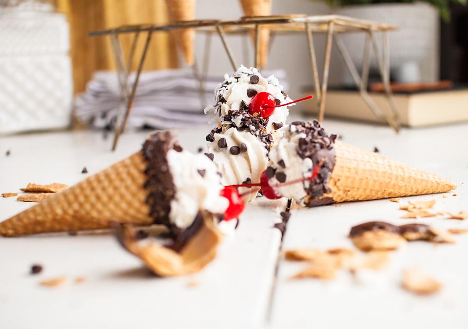 Two ice cream cones with whipped cream, chocolate chips, and cherries have fallen on a white surface, with broken cone pieces scattered around.