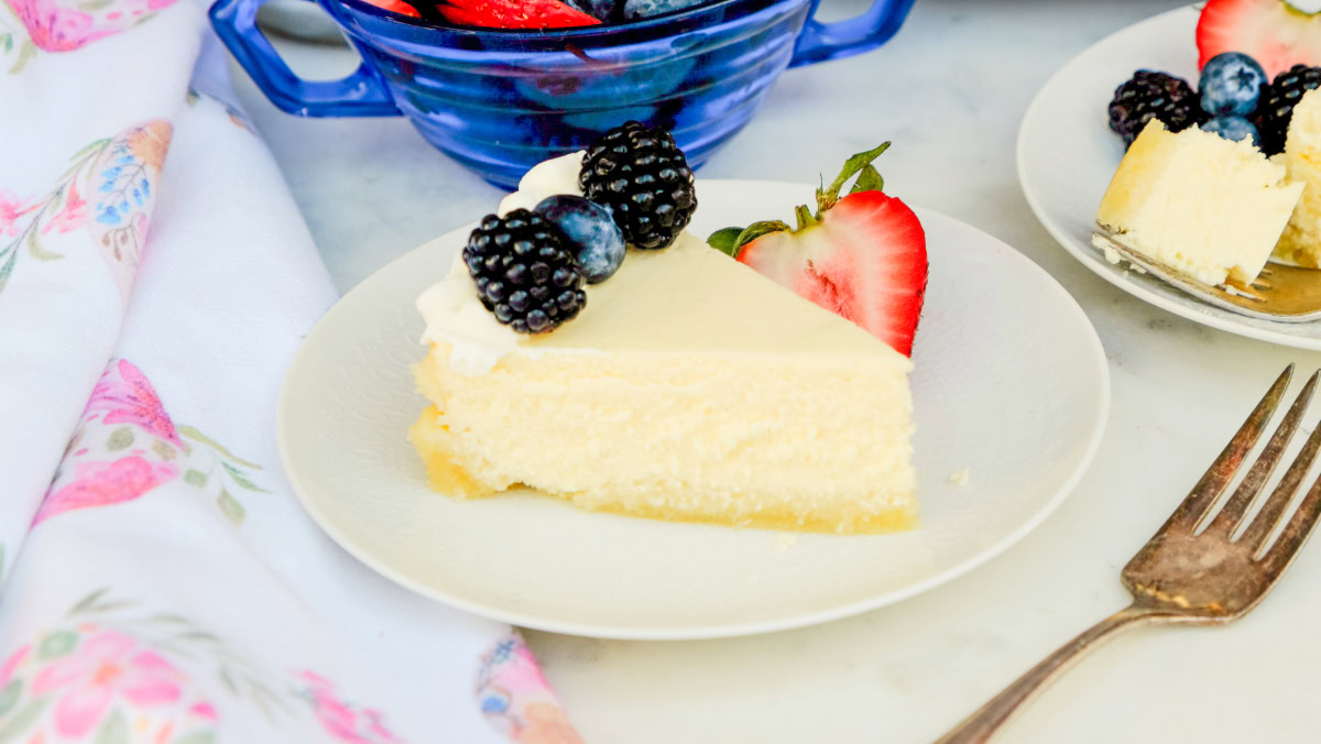 A slice of cheesecake topped with blackberries, a blueberry, and a strawberry half on a white plate, with a fork and a bowl of mixed berries nearby.