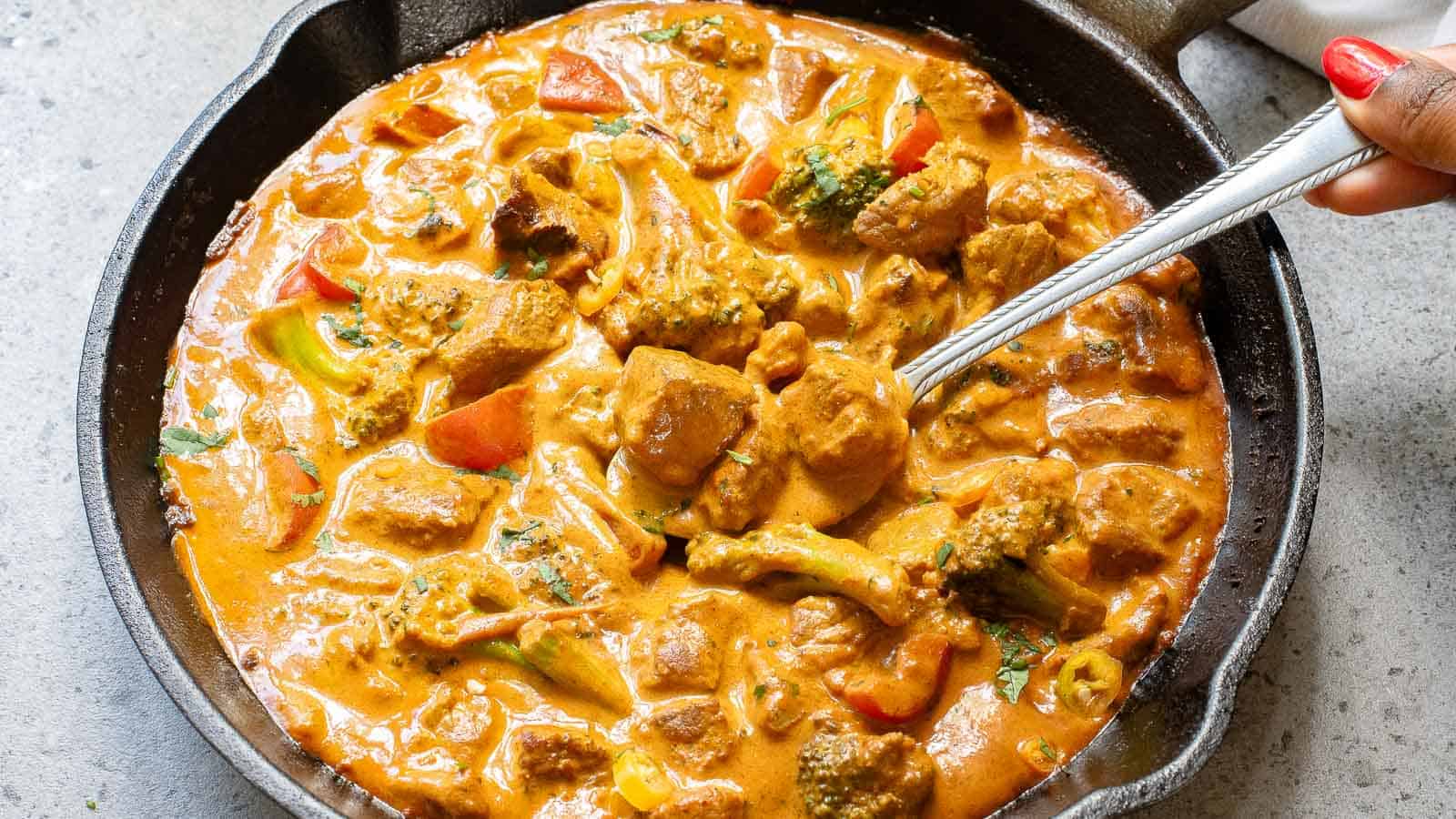 A cast iron skillet filled with creamy orange curry containing chunks of meat and vegetables, with a spoon scooping some from the dish.