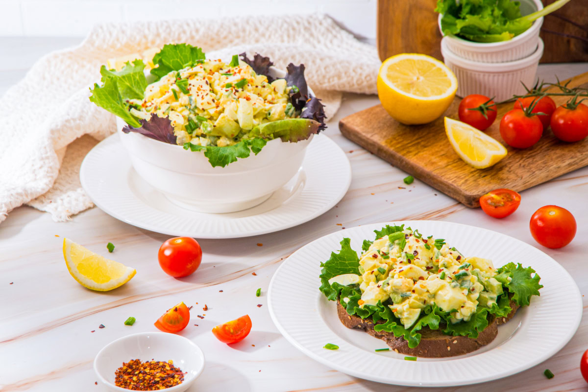 A bowl and a slice of bread topped with egg salad on lettuce, surrounded by cherry tomatoes, lemon wedges, and seasoning on a white surface.