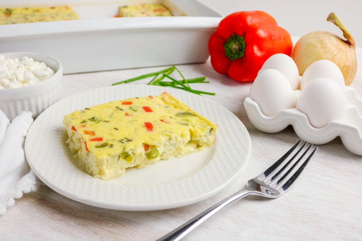 A slice of vegetable egg casserole on a white plate with a fork, next to eggs, red bell pepper, onion, cottage cheese, and chives on a white table.