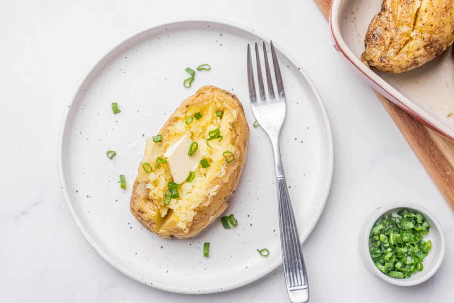 A baked potato topped with butter and chopped green onions on a white plate with a fork, next to a small bowl of green onions.