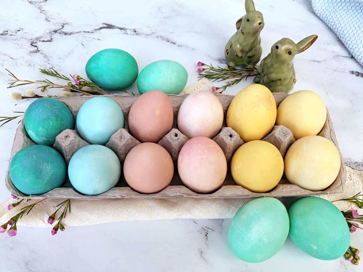 A carton of pastel-colored Easter eggs sits on a marble surface with small flowers and two ceramic rabbit figurines in the background.