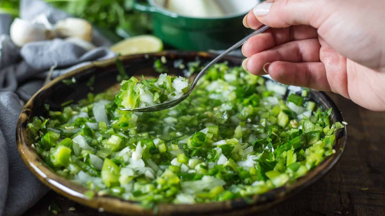 A hand holds a spoon over a bowl of finely chopped green onions, cilantro, and white onions mixed in liquid, with garlic and lemon in the background.
