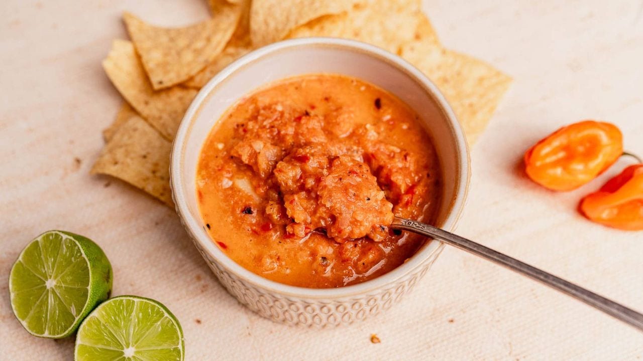 A bowl of orange-red salsa with a spoon, surrounded by tortilla chips, lime halves, and a whole habanero pepper on a light surface.