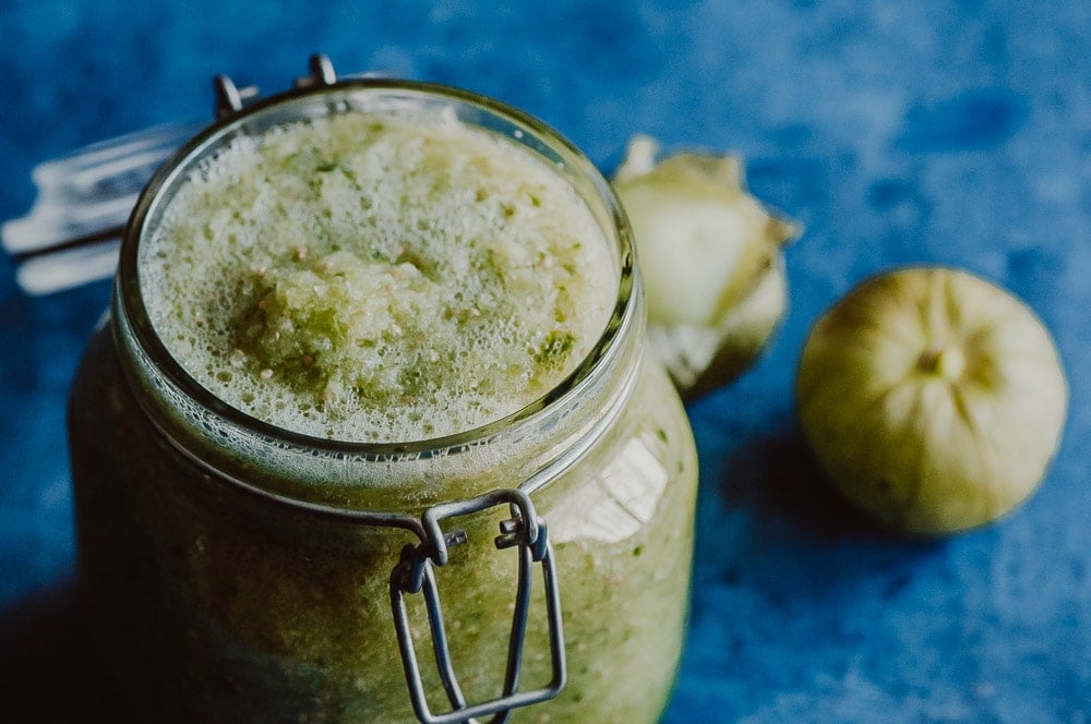 A glass jar filled with green salsa, with a foamy surface, sits on a blue background next to two whole tomatillos.