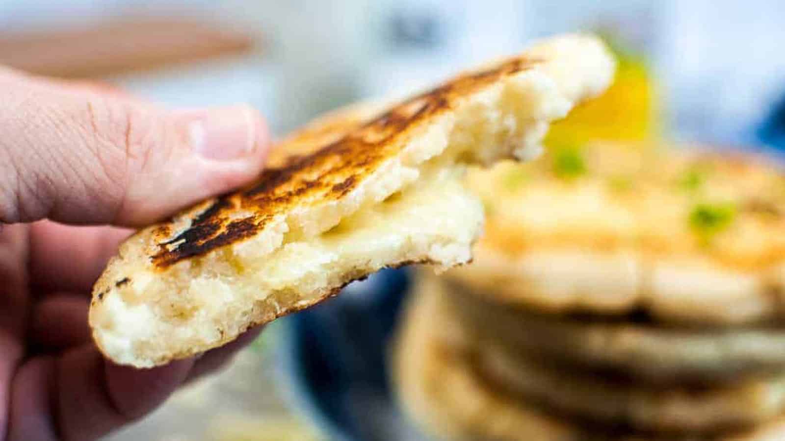 A hand holds a half-eaten, grilled arepa showing melted cheese inside, with a stack of arepas blurred in the background.