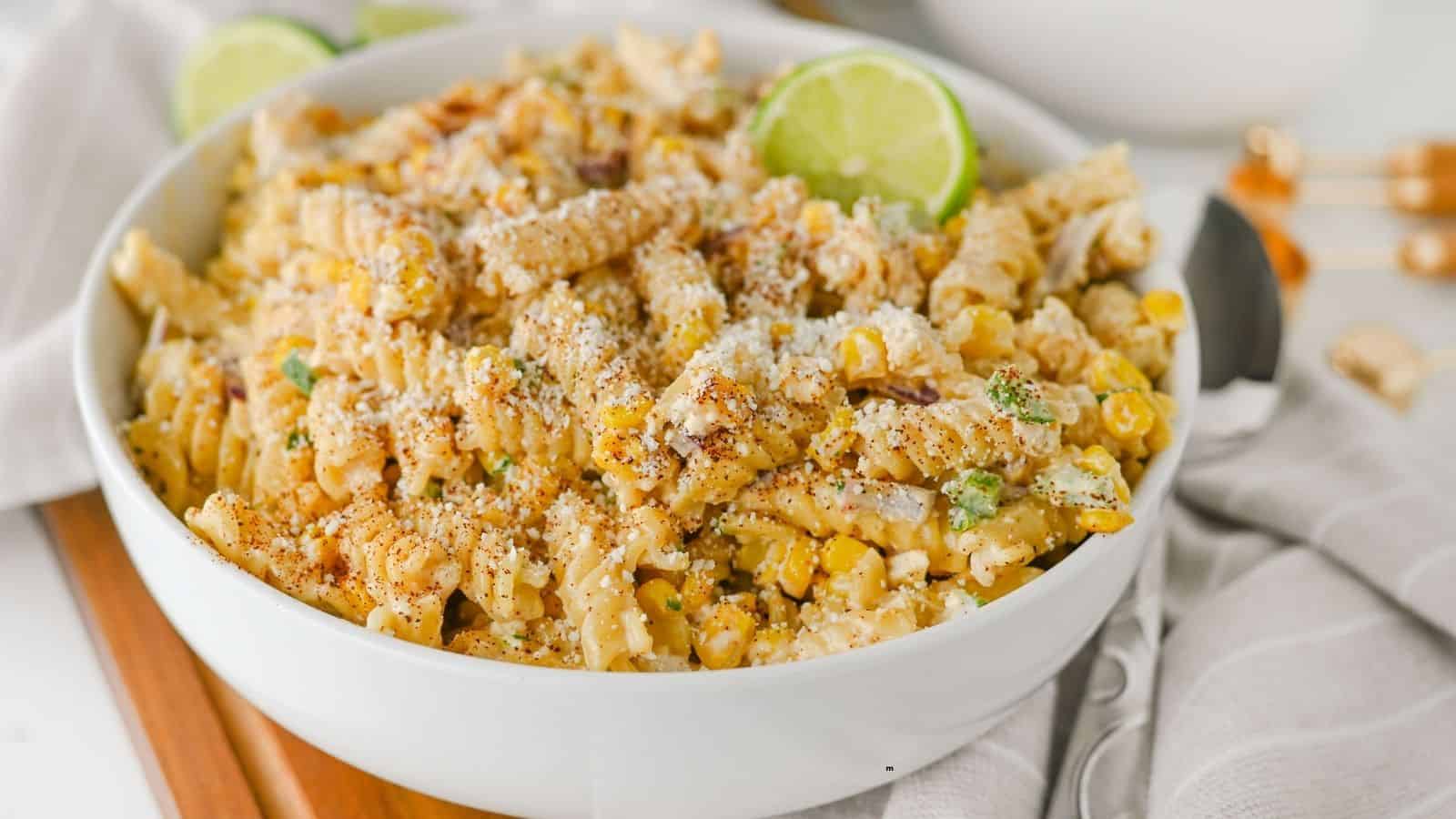 A white bowl filled with creamy rotini pasta mixed with corn, green herbs, grated cheese, and black pepper, garnished with lime slices.