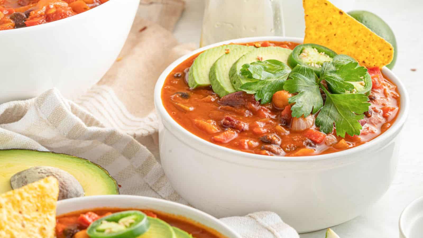 A white bowl of chili topped with sliced avocado, jalapeño, cilantro, and a tortilla chip is placed on a striped cloth next to a halved avocado.