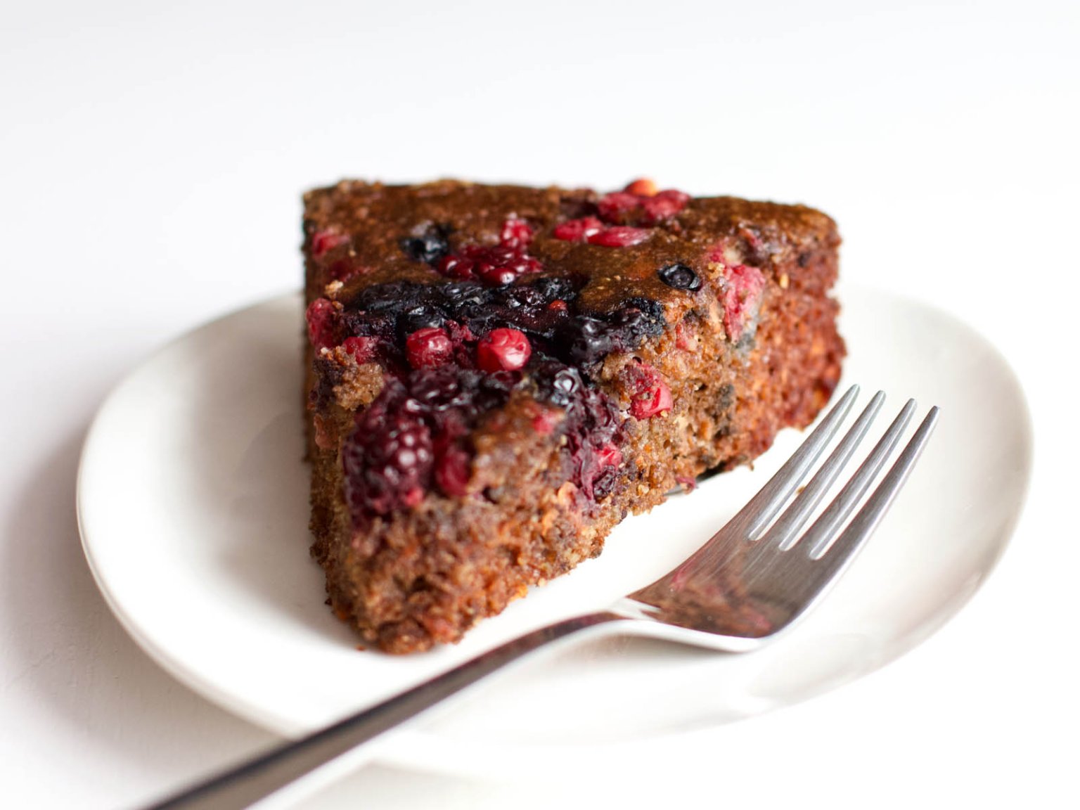 A slice of dark berry cake with mixed berries on top, served on a white plate with a fork.