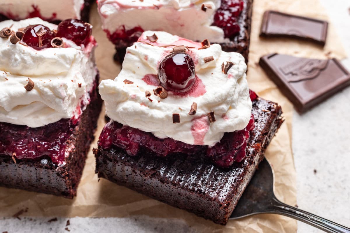 A piece of chocolate cake topped with cherry filling, whipped cream, a cherry, and chocolate shavings on a spatula, with chocolate pieces in the background.