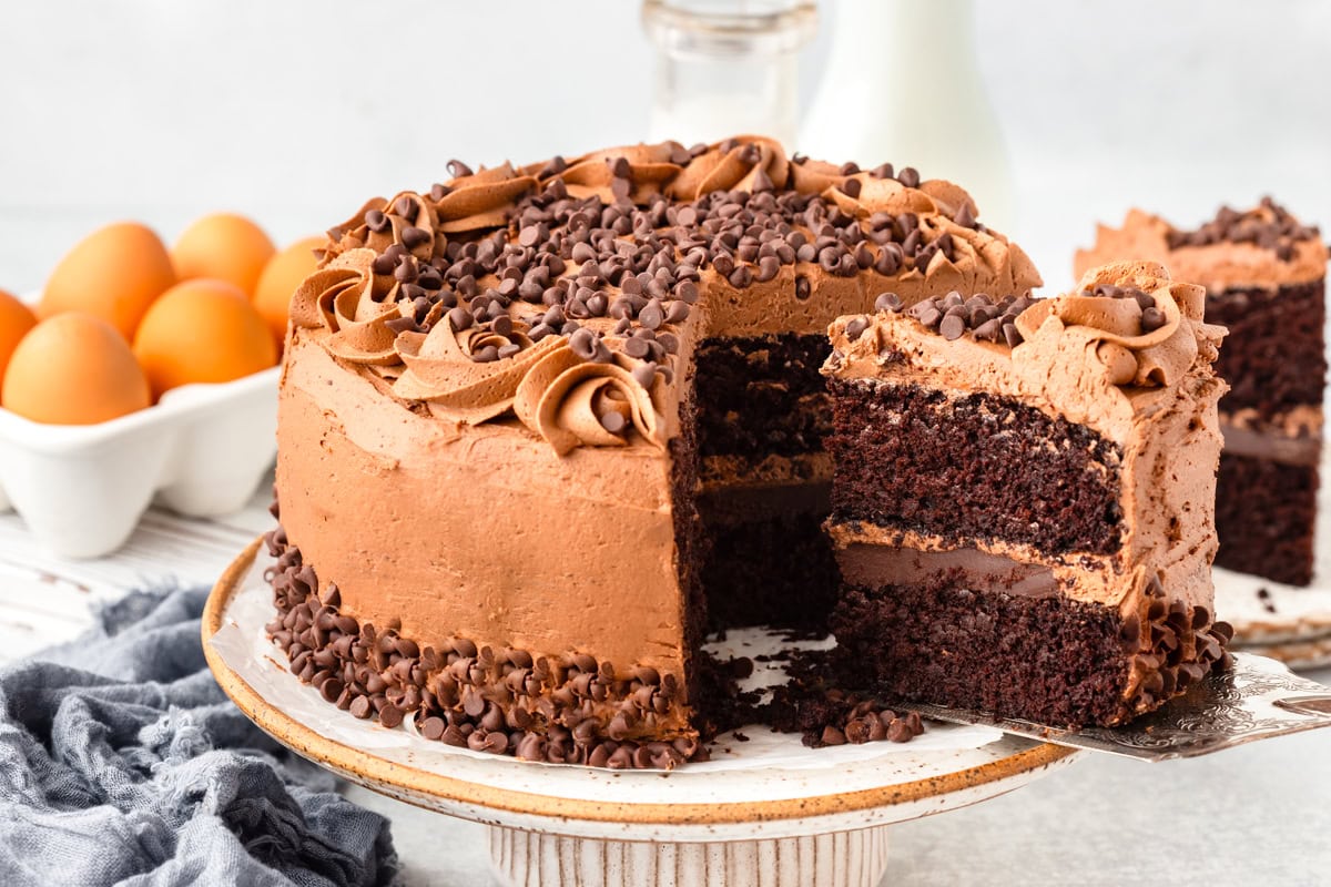 A chocolate layer cake with chocolate frosting and chocolate chips sits on a cake stand; a slice is being served. Eggs, milk, and a cloth are in the background.