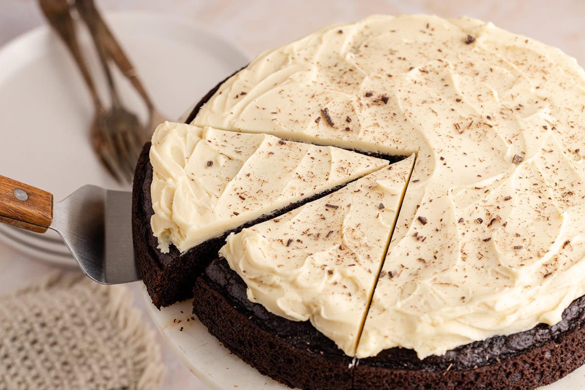 A sliced chocolate cake with white frosting and chocolate shavings sits on a cake stand; a server lifts one piece. Plates and forks are in the background.