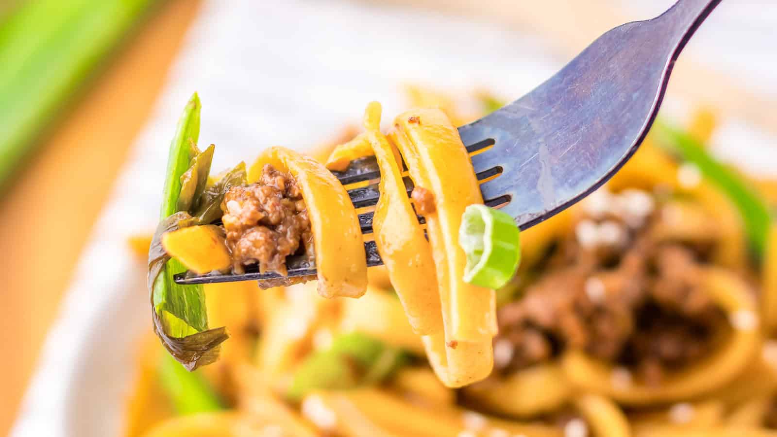 A close-up of a fork holding noodles with ground meat, green onions, and sauce.