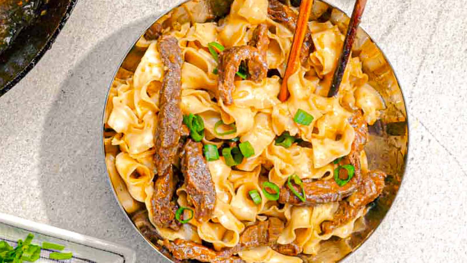 A bowl of wide, ribbon-like noodles mixed with sliced beef and topped with chopped green onions, with chopsticks picking up some noodles.